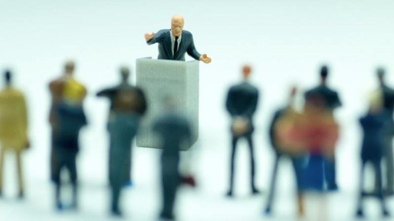 Cartoon figures of busines people looking at a man speaking from a podium.